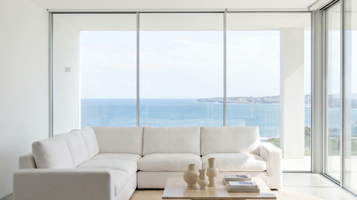 Sala blanca moderna con sofá de lino, mesa de travertino y vista panorámica al mar a través de ventanales de piso a techo