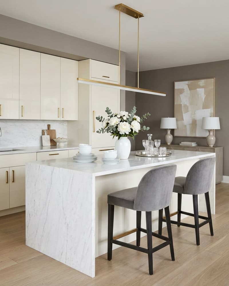 Cocina estilo hotel boutique con gabinetes crema y tiradores brass, isla de mármol con waterfall, peonías blancas, lámpara brass lineal y arte abstracto